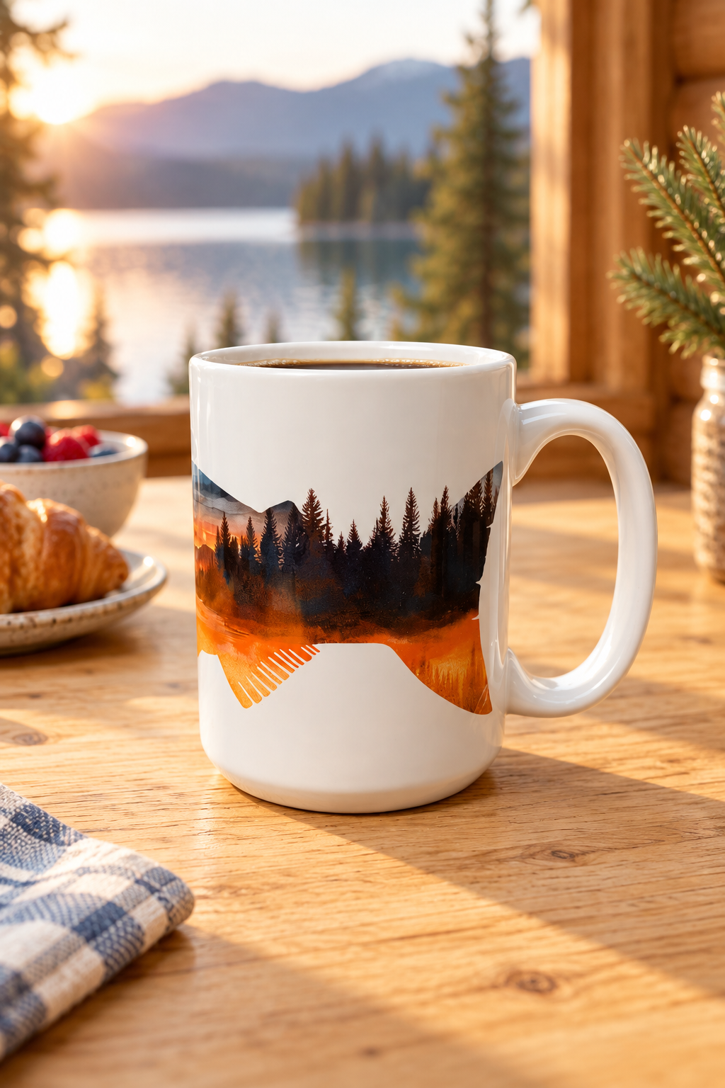 A white ceramic coffee mug with a scenic mountain and forest landscape print, filled with coffee and set on a wooden table in a cozy cafe setting with a lake and mountains in the background.